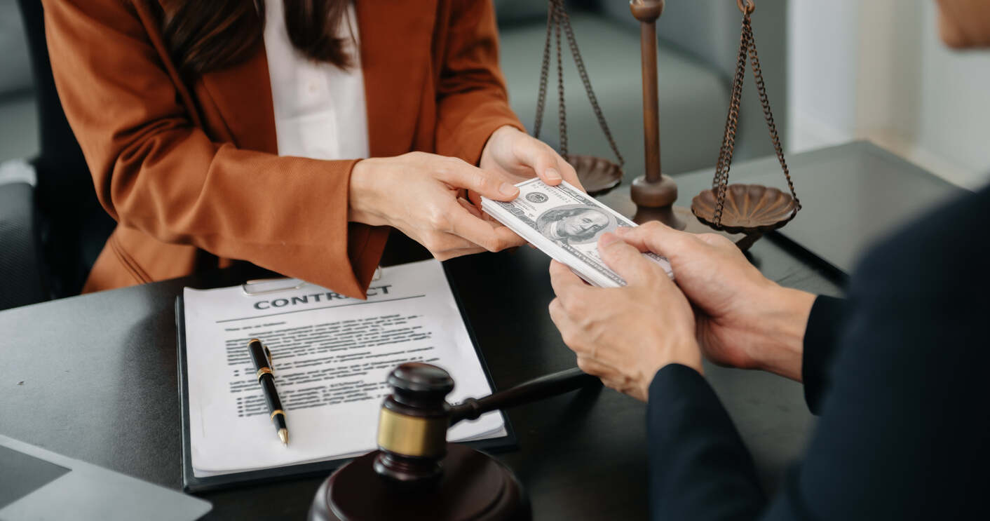 A legal professional handing money to a client over a contract with a gavel and scales of justice in the background, symbolizing legal negotiations or settlements.
