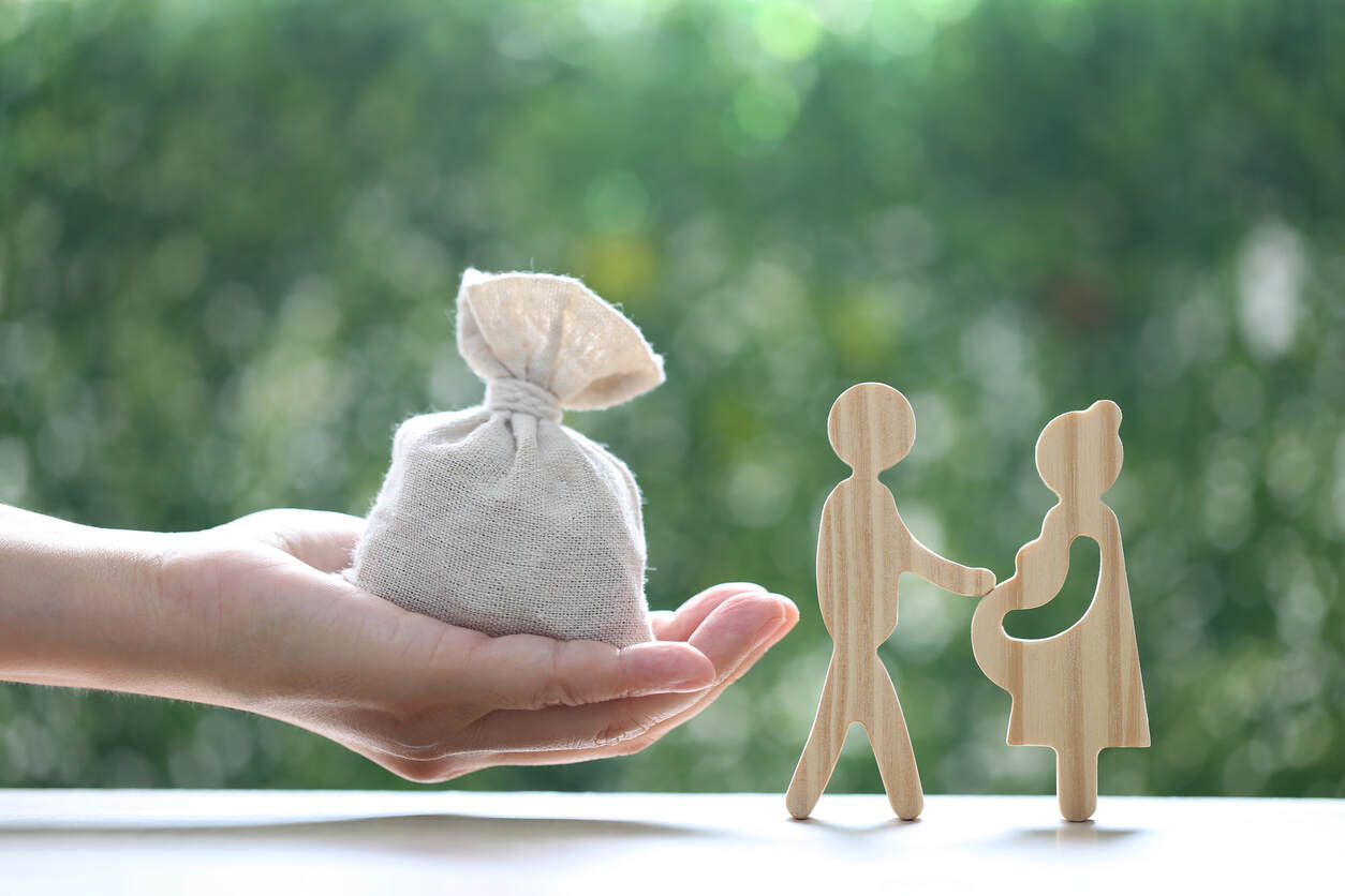 A hand holding a bag of money, with wooden figurines of a man and a pregnant woman symbolizing child custody and visitation rights.